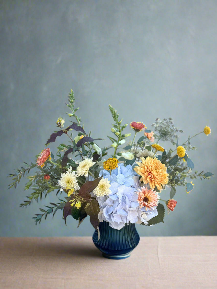 Bouquet of flowers in a blue vase on a wooden surface with a white brick wall background