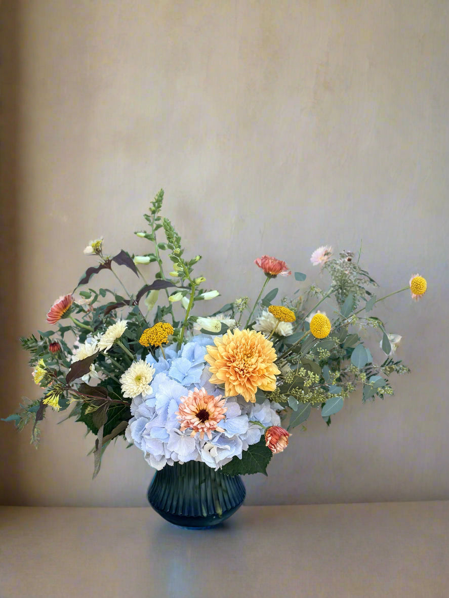 Floral arrangement in a blue vase on a wooden table with a light blue brick wall background