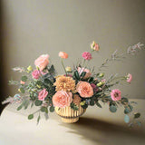 A floral arrangement with peach, pink, and soft yellow blooms in a matte gold vase, displayed on a clear stand.