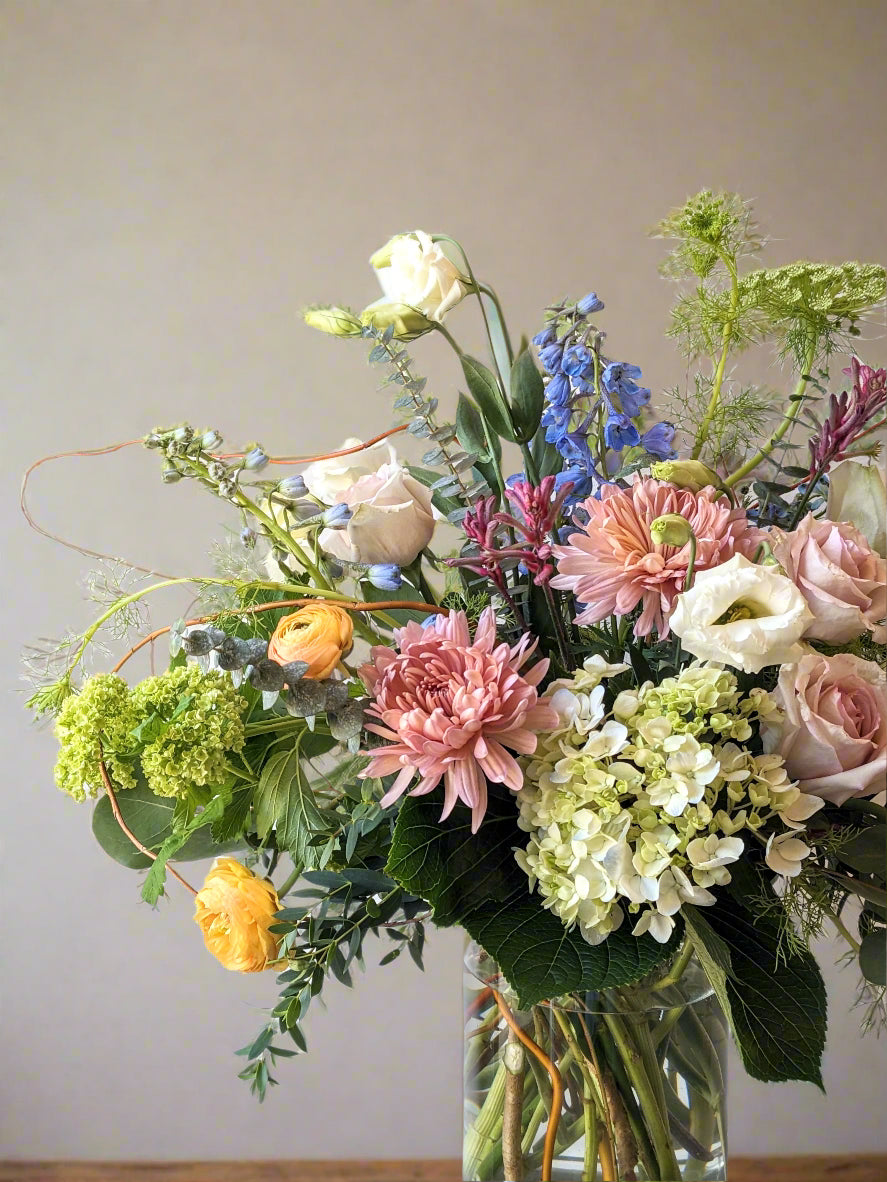 A vibrant floral arrangement featuring a variety of flowers including roses and hydrangea, with green branches and textural greenery, arranged in a clear glass vase.