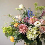 A vibrant floral arrangement featuring a variety of flowers including roses and hydrangea, with green branches and textural greenery, arranged in a clear glass vase.