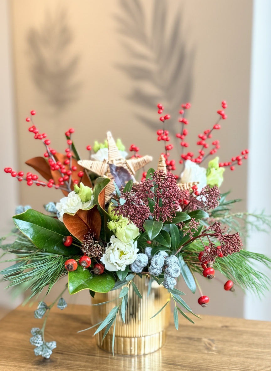 Decorative floral arrangement with red berries and green leaves in a gold vase on a wooden surface.