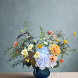Bouquet of flowers in a blue vase on a wooden surface with a white brick wall background