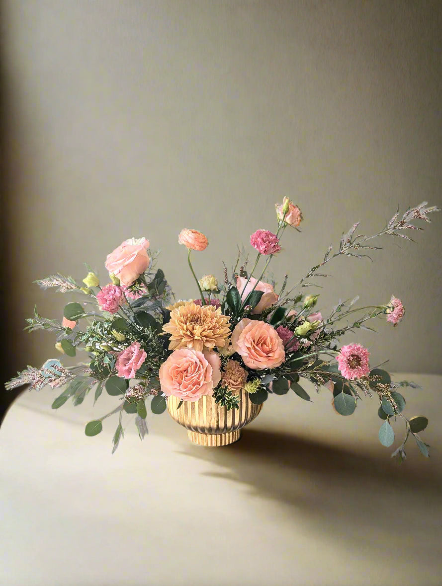 A floral arrangement with peach, pink, and soft yellow blooms in a matte gold vase, displayed on a clear stand.