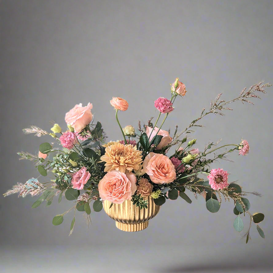 A floral arrangement with peach, pink, and soft yellow blooms in a matte gold vase, displayed on a clear stand.